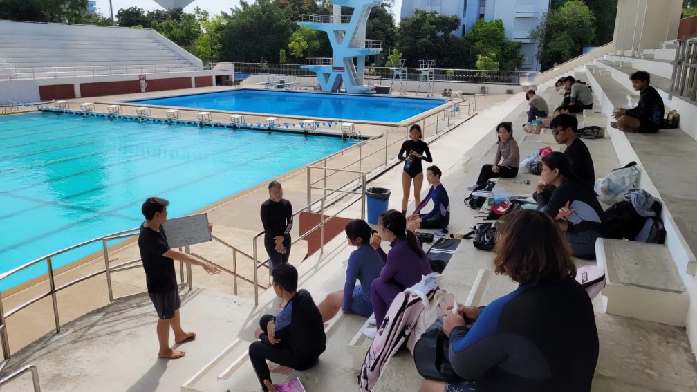 A group of freediver gathered at a swimming pool, some sitting, others standing. The instructor in the foreground are giving a briefing or instruction, holding a clipboard. The attendees are dressed in casual and swimwear. The pool is Olympic-sized with clear blue water, and there's a diving platform in the background under a bright, sunny sky