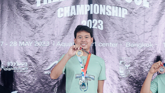 A person stands on a podium at the AIDA Thailand Freediving Pool Championship 2023, smiling and holding a gold medal. They are wearing a light green t-shirt with event logos and text. The background is a crinkled banner with event details, including the date and location in Bangkok. The left side of the image is slightly obscured by another individual's arm, also holding a medal.